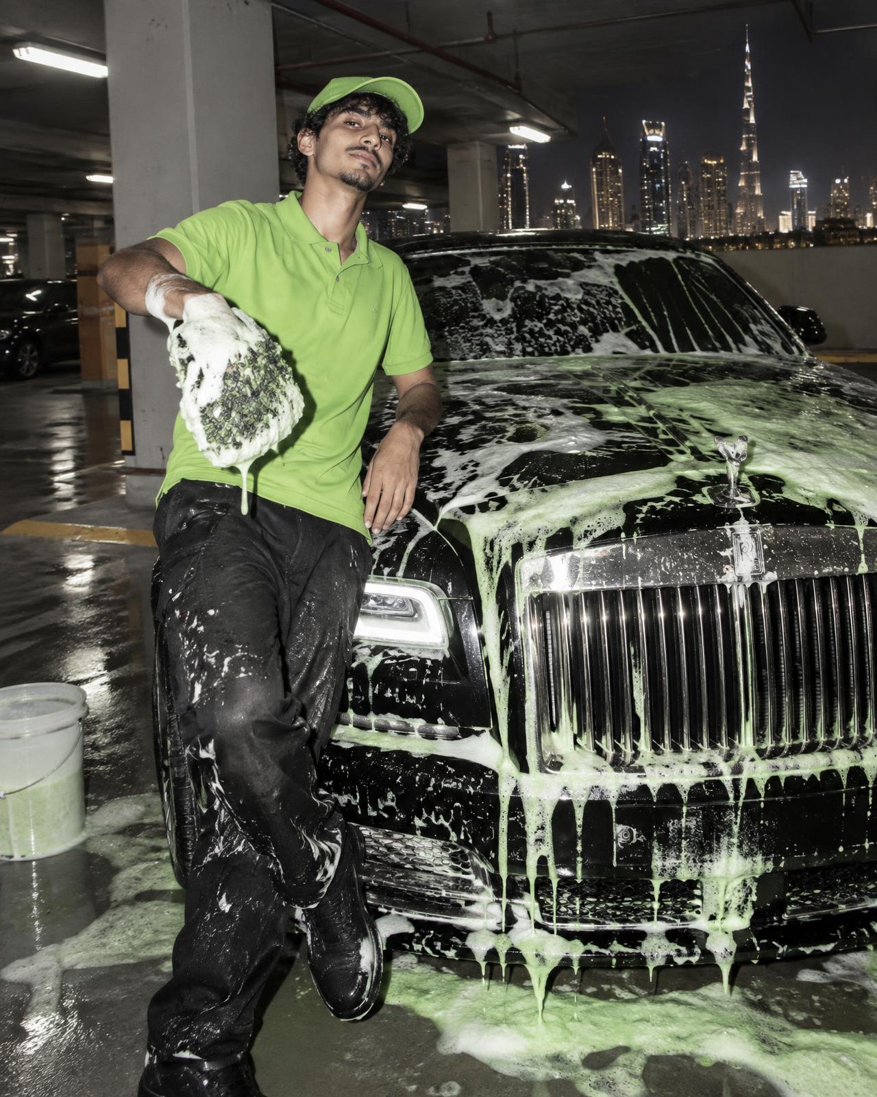 Motor Ritual tech in lime polo leaning on a foam-covered luxury car in a Dubai parking garage at night